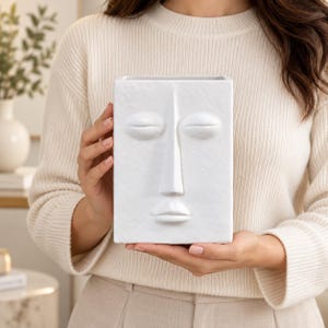 Puede incluir: Un jarrón rectangular de cerámica blanca que presenta un rostro humano abstracto esculpido en la superficie frontal. El diseño incluye ojos cerrados, una nariz prominente y labios definidos, todo en un acabado blanco mate con textura de piedra.
