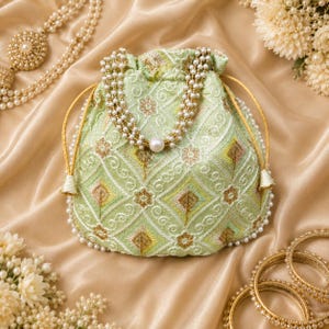 May include: A light green drawstring pouch featuring intricate white and gold embroidery in a geometric diamond pattern. The bag is adorned with a pearl-accented beaded handle and a border of small white beads along the bottom edge.