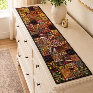 May include: A long, rectangular table runner featuring a patchwork design composed of numerous small, square fabric panels. Each panel displays a unique combination of dark, rich colours, intricate metallic embroidery, sequins, and varied textures.