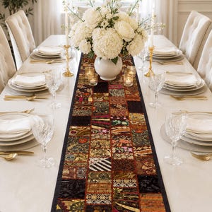 May include: A long rectangular table runner featuring a patchwork design of various dark-toned fabric squares with intricate gold, red, and black embroidery, metallic thread accents, and decorative patterns, centred on a white dining table.