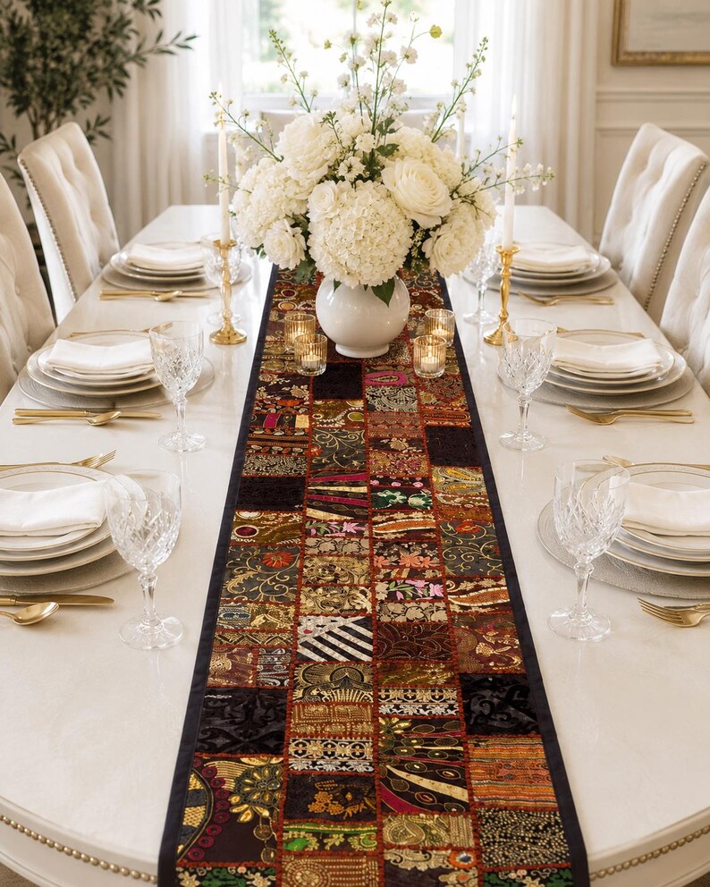 May include: A long rectangular table runner featuring a patchwork design of various dark-toned fabric squares with intricate gold, red, and black embroidery, metallic thread accents, and decorative patterns, centred on a white dining table.
