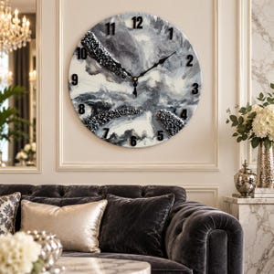 May include: A round wall clock featuring a marbled black, white, and gray resin face with black numerals and three black hands. The clock face includes textured, crystalline accents in dark gray and black, creating a layered, abstract appearance.