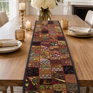 May include: A rectangular table runner featuring a patchwork design of various fabric squares in deep red, gold, black, and green tones. The runner displays intricate embroidery, metallic thread accents, and small reflective mirror embellishments.