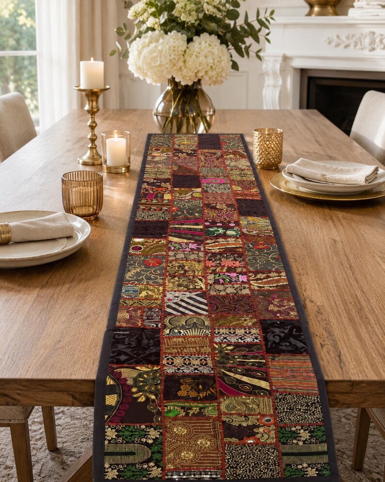 May include: A rectangular table runner featuring a patchwork design of various fabric squares in deep red, gold, black, and green tones. The runner displays intricate embroidery, metallic thread accents, and small reflective mirror embellishments.