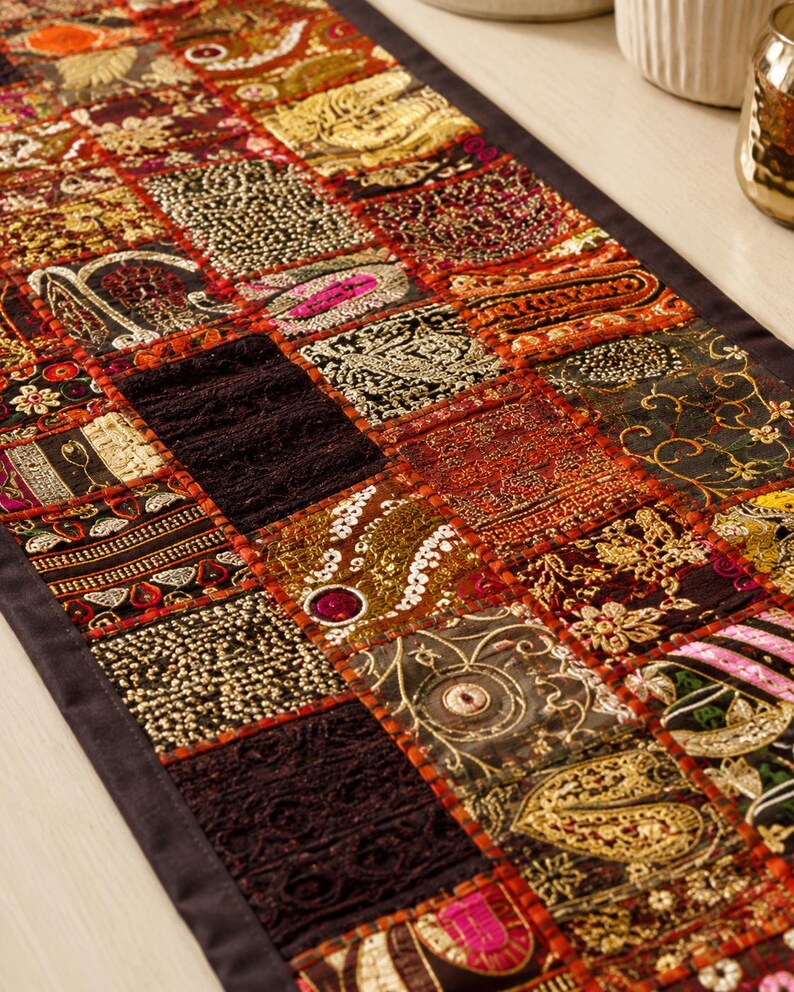 May include: A rectangular table runner featuring a patchwork design of various fabric squares in shades of red, gold, black, and orange. The squares are decorated with intricate metallic embroidery, sequins, beads, and textured lace patterns.