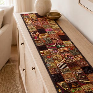 May include: A long, rectangular table runner featuring a patchwork design composed of numerous small, square fabric panels. Each panel displays distinct, intricate embroidery, metallic thread accents, and varied patterns in dark, rich, and vibrant colours.