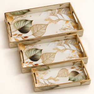 May include: A set of three rectangular serving trays with integrated handles, featuring a cream-colored base decorated with gold-lined leaf illustrations, soft green shaded foliage, and white leaf accents, arranged in a nested formation.