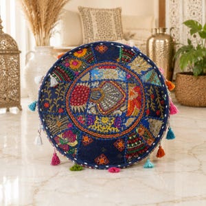 May include: A round floor cushion featuring a dark blue fabric base with a patchwork design of embroidered floral and geometric patterns in vibrant colors. The cushion is accented with small mirrors, metallic thread, and colorful tassels along the edge.