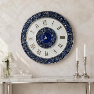 Può includere: Un orologio da parete rotondo con quadrante bianco e numeri romani neri, circondato da un'ampia cornice di pietre blu e argento frantumate. Il centro dell'orologio presenta un inserto circolare blu intenso e lancette in metallo lavorato.