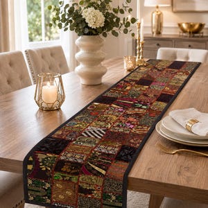 May include: A long rectangular table runner featuring a patchwork design of various dark-toned fabric squares with intricate gold, red, green, and black patterns. The runner has a solid black border and is laid flat across a wooden dining table.