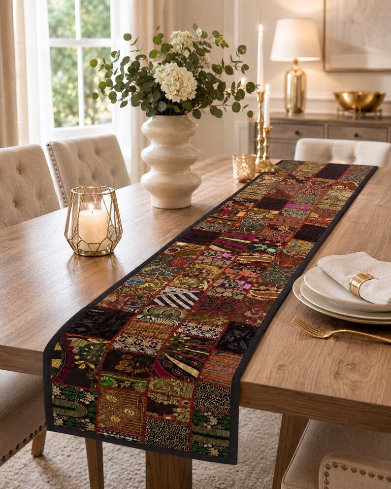 May include: A long rectangular table runner featuring a patchwork design of various dark-toned fabric squares with intricate gold, red, green, and black patterns. The runner has a solid black border and is laid flat across a wooden dining table.