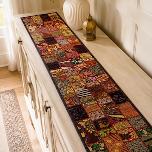 May include: A long rectangular table runner featuring a patchwork design of various fabric squares in deep red, gold, green, and black tones. The runner is decorated with intricate embroidery, metallic thread accents, and small circular mirrors.