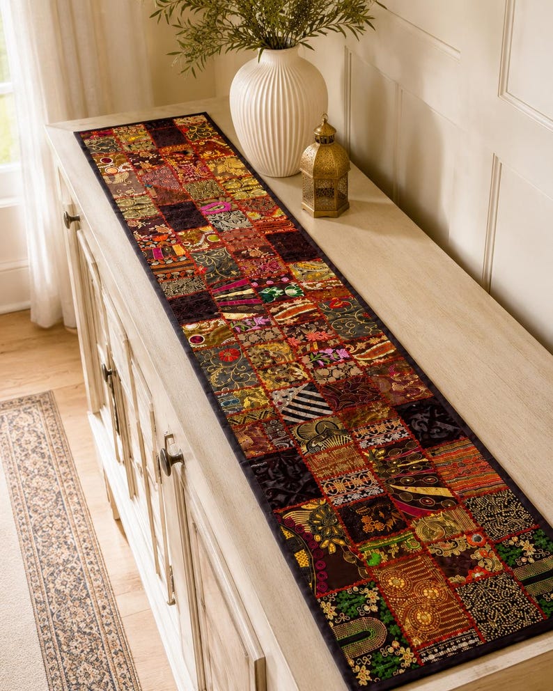 May include: A long rectangular table runner featuring a patchwork design of various fabric squares in deep red, gold, green, and black tones. The runner is decorated with intricate embroidery, metallic thread accents, and small circular mirrors.