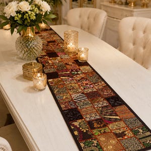 May include: A long rectangular table runner featuring a patchwork design of various dark-toned fabric squares. Each square displays intricate embroidery, metallic thread accents, and small mirror embellishments in shades of gold, red, and black.