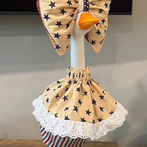May include: A decorative ceramic goose figurine wearing a tan fabric outfit with navy blue stars. The goose features a large matching bow on its head, a skirt with a white eyelet lace trim, and a red, white, and blue striped fabric base.