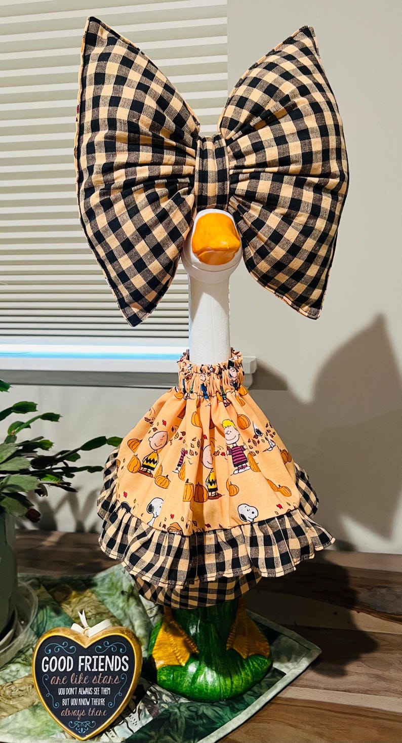 May include: A white goose figurine wearing a large black and tan buffalo check bow on its head and a peach-colored skirt featuring a Peanuts character print with pumpkins and leaves, finished with a matching buffalo check ruffled hem trim.