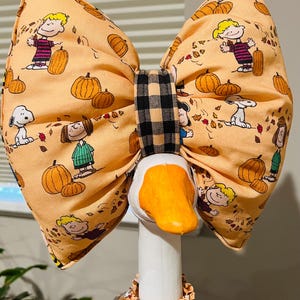May include: A large fabric bow featuring a Peanuts character print with Snoopy, Charlie Brown, and pumpkins on an orange background. The bow has a black and tan buffalo check center knot and is positioned on the head of a white decorative goose.