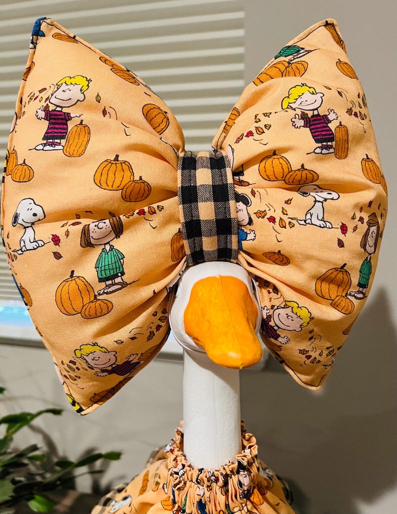 May include: A large fabric bow featuring a Peanuts character print with Snoopy, Charlie Brown, and pumpkins on an orange background. The bow has a black and tan buffalo check center knot and is positioned on the head of a white decorative goose.