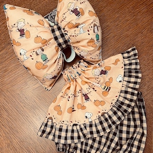 May include: A large fabric hair bow and a matching ruffled skirt featuring a light orange background with a repeating pattern of Peanuts characters and pumpkins, accented by a black and white buffalo check fabric on the bow center and skirt trim.