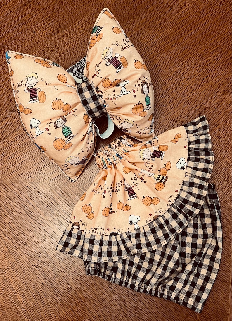 May include: A large fabric hair bow and a matching ruffled skirt featuring a light orange background with a repeating pattern of Peanuts characters and pumpkins, accented by a black and white buffalo check fabric on the bow center and skirt trim.