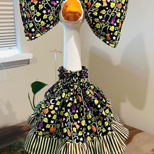 May include: A white goose statue wearing a black fabric outfit with a Halloween print featuring orange pumpkins, purple bats, and brooms. The outfit includes a large bow headpiece and a ruffled skirt with a green and brown striped trim at the base.