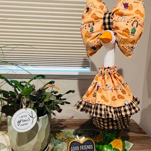 May include: A decorative goose figurine wearing a peach-colored fabric outfit featuring Peanuts characters and pumpkins, accented with a black and tan buffalo plaid trim on the skirt and a matching oversized bow on its head.