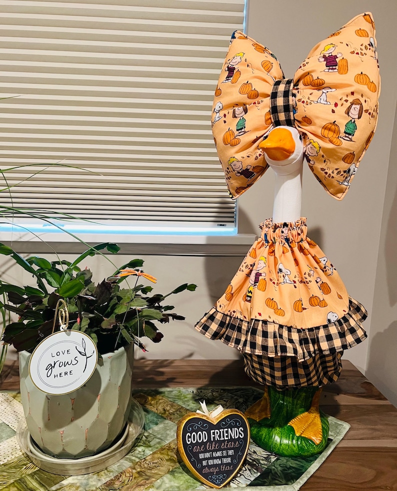 May include: A decorative goose figurine wearing a peach-colored fabric outfit featuring Peanuts characters and pumpkins, accented with a black and tan buffalo plaid trim on the skirt and a matching oversized bow on its head.