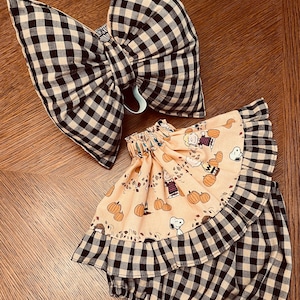 May include: A large hair bow and a ruffled skirt featuring a black and white gingham check pattern. The skirt includes a peach-colored fabric panel printed with Snoopy, Charlie Brown, and pumpkin illustrations, finished with a gathered elastic waist.