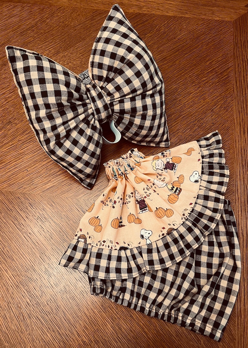 May include: A large hair bow and a ruffled skirt featuring a black and white gingham check pattern. The skirt includes a peach-colored fabric panel printed with Snoopy, Charlie Brown, and pumpkin illustrations, finished with a gathered elastic waist.