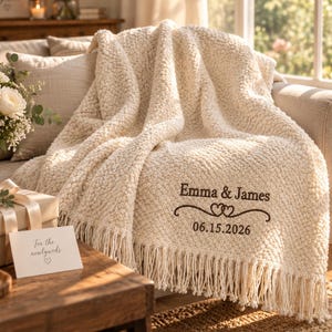 May include: A cream-coloured, textured knit throw blanket with fringe trim. The blanket features dark brown embroidery that reads "Emma & James" above two interlocking heart icons and the date "06.15.2026" centred on the lower corner.