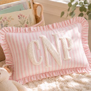 May include: A rectangular throw pillow featuring a light pink and white vertical striped pattern with a ruffled border. The center of the pillow displays the large, textured, cream-colored embroidered monogram letters CNP in a serif font.