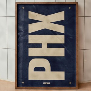 Könnte beinhalten: Ein gerahmtes vertikales Poster mit einem dunkelblauen, texturierten Hintergrund und den großen, cremefarbenen Buchstaben PHX, die vertikal gestapelt sind. Das Wort ARIZONA steht in kleinerer cremefarbener Schrift unten, mit vier kleinen Pluszeichen.