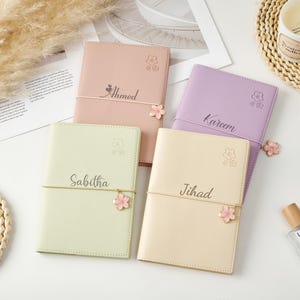 May include: Four rectangular notebooks in pastel shades of pink, lavender, light green, and cream. Each notebook features a personalised name, a small embossed flower design, and a thin elastic band with a dangling pink flower charm attached.