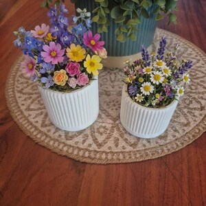 Peut inclure: Deux petits pots en céramique blanche cannelée contenant des compositions florales artificielles. Le pot de gauche présente des fleurs roses, jaunes et bleues. Le pot de droite contient des marguerites blanches, de la lavande et des petites fleurs.