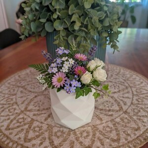 Peut inclure: Un petit vase géométrique blanc contenant un arrangement floral composé de marguerites roses, de myosotis bleu clair, de roses blanches, de brins de lavande et de feuilles de lierre vert. Le vase présente une surface facettée et angulaire.