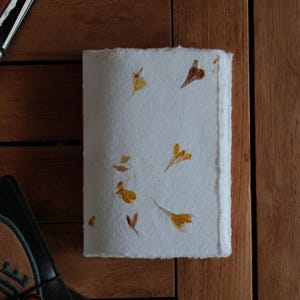 May include: A rectangular notebook with a white, textured, handmade paper cover embedded with small, dried yellow and brown flower petals. The paper has rough, deckled edges, and the notebook is positioned vertically on a wooden surface.