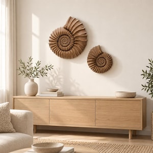 May include: Two brown, spiral-shaped ammonite fossil wall sculptures mounted on a plain wall above a long, light-toned wooden sideboard. The larger sculpture is positioned to the left, while the smaller one is placed to the right.