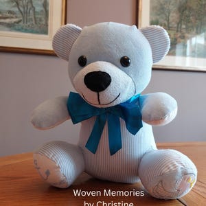 May include: A light blue teddy bear made from striped fabric with a dark blue satin bow around its neck. The bear features black plastic eyes, a black nose, and embroidered details on the feet, including an anchor and a bird. Text reads Woven Memories by Christine.