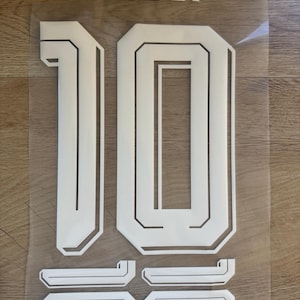 May include: A white heat transfer vinyl decal set featuring the name MESSI in block lettering, a large number 10, and two smaller versions of the number 10, all presented on a clear adhesive backing sheet for application onto sports apparel.