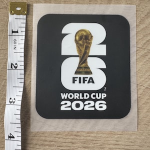 Peut inclure: Un autocollant noir carr&eacute; avec le texte blanc 26 et le logo de la Coupe du Monde de la FIFA 2026 incluant un troph&eacute;e dor&eacute;. Un ruban &agrave; mesurer blanc est plac&eacute; &agrave; gauche, indiquant une hauteur d'autocollant d'environ 11,5 cm.