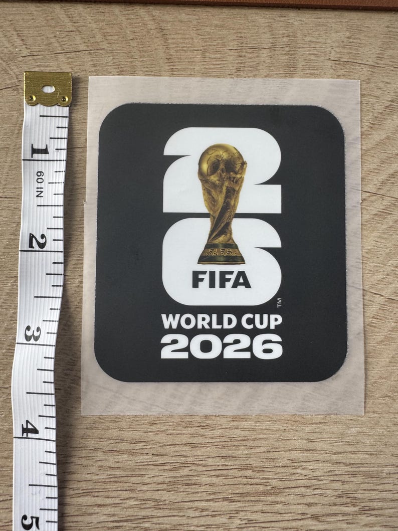 Peut inclure: Un autocollant noir carr&eacute; avec le texte blanc 26 et le logo de la Coupe du Monde de la FIFA 2026 incluant un troph&eacute;e dor&eacute;. Un ruban &agrave; mesurer blanc est plac&eacute; &agrave; gauche, indiquant une hauteur d'autocollant d'environ 11,5 cm.