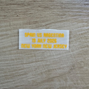 May include: A rectangular white iron-on patch featuring yellow embroidered text that reads: SPAIN US ARGENTINA, 19 JULY 2026, NEW YORK NEW JERSEY. The patch has a clean, block-style font and is designed for application onto fabric surfaces.