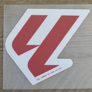 May include: A die-cut vinyl decal featuring a large, stylized red letter LL logo on a white background. The design includes the text THE POWER OF OUR FÚTBOL printed in small red capital letters at the bottom right of the graphic element.
