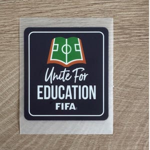 Peut inclure: Un autocollant carr&eacute; aux coins arrondis avec un fond bleu fonc&eacute; et une bordure blanche. Le motif repr&eacute;sente un livre vert ouvert avec des lignes de terrain de football blanches, au-dessus du texte Unite For EDUCATION et du logo FIFA en blanc.