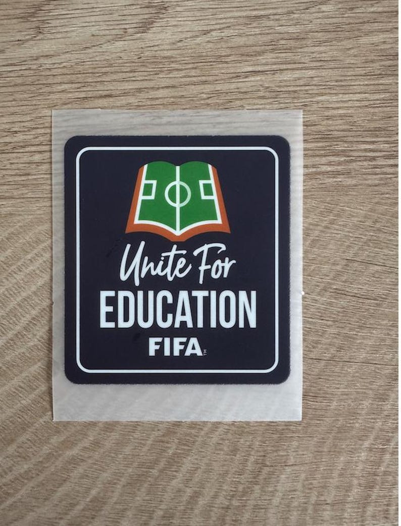 Peut inclure: Un autocollant carr&eacute; aux coins arrondis avec un fond bleu fonc&eacute; et une bordure blanche. Le motif repr&eacute;sente un livre vert ouvert avec des lignes de terrain de football blanches, au-dessus du texte Unite For EDUCATION et du logo FIFA en blanc.