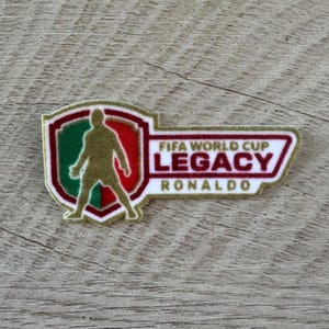 May include: A rectangular patch with a shield-shaped left side featuring a gold silhouette of a soccer player against a green and red background. The right side contains the text FIFA WORLD CUP LEGACY RONALDO in dark red and gold lettering.