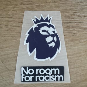 May include: A vinyl decal featuring a dark blue lion head icon with a crown on top, positioned above a rectangular black label with the white text "No room FOR racism" printed in a bold, sans-serif font on a clear adhesive backing.