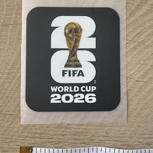 Peut inclure: Un autocollant carr&eacute; noir avec le texte blanc 26, un troph&eacute;e dor&eacute; de la Coupe du Monde de la FIFA &agrave; l'int&eacute;rieur du z&eacute;ro, et le texte FIFA WORLD CUP 2026 en dessous. Un ruban &agrave; mesurer blanc indique une largeur d'environ 7,6 cm.