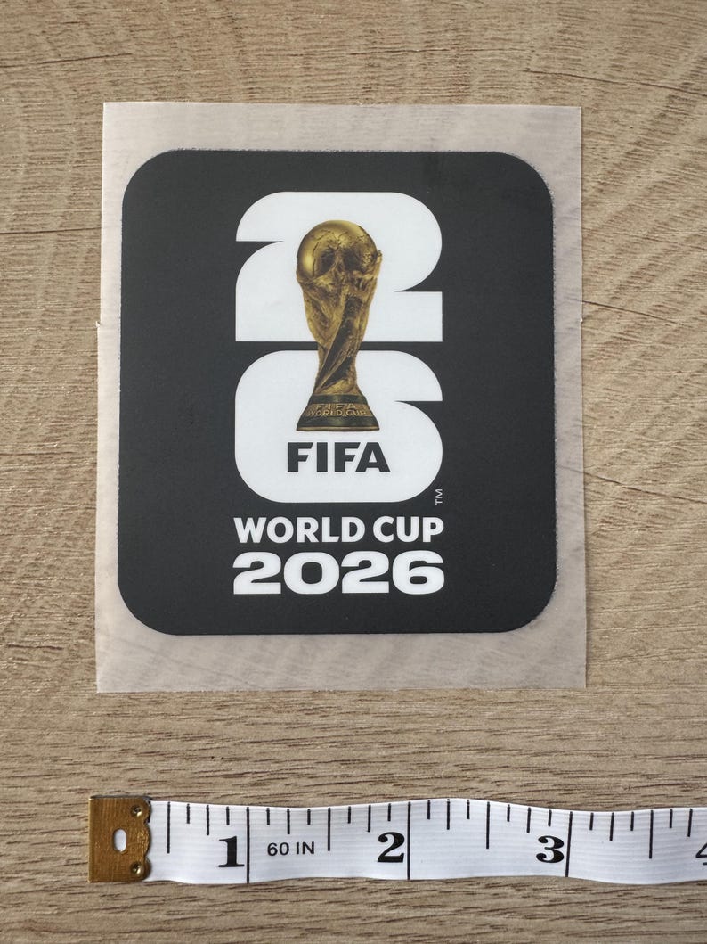 Peut inclure: Un autocollant carr&eacute; noir avec le texte blanc 26, un troph&eacute;e dor&eacute; de la Coupe du Monde de la FIFA &agrave; l'int&eacute;rieur du z&eacute;ro, et le texte FIFA WORLD CUP 2026 en dessous. Un ruban &agrave; mesurer blanc indique une largeur d'environ 7,6 cm.