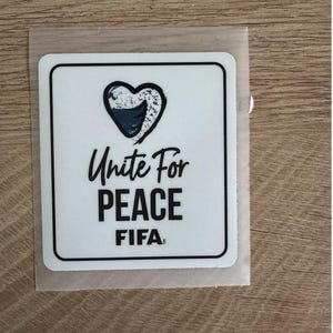 Peut inclure: Un autocollant carr&eacute; blanc aux coins arrondis avec un contour noir. Le motif comprend un c&oelig;ur avec un motif textur&eacute; bleu et blanc, suivi du texte noir Unite For PEACE et du mot FIFA &eacute;crit dans une police de caract&egrave;res plus petite.